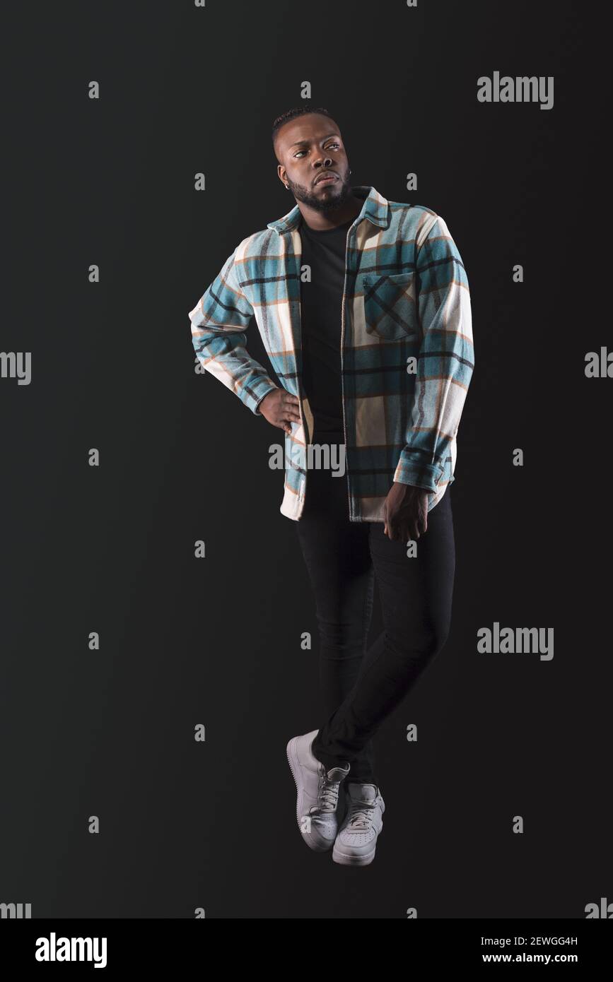Schöner schwarzer Mann, der ernst zur Seite steht, trägt blaues kariertes Hemd und Jeans, Beine gekreuzt. Ganzkörper. Blauer Hintergrund. Stockfoto