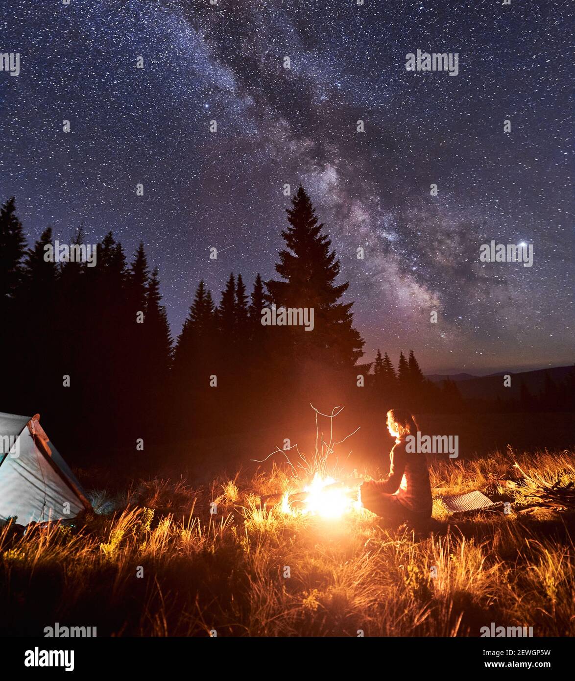 Seitenansicht der weiblichen Touristen, die sich am Lagerfeuer auf dem Hintergrund des Zeltes und des Fichtenwaldes unter dem Sternenhimmel ausruhen, auf dem die Milchstraße sichtbar ist. Lagerfeuer mit einer großen Flamme unter dem Nachthimmel Stockfoto