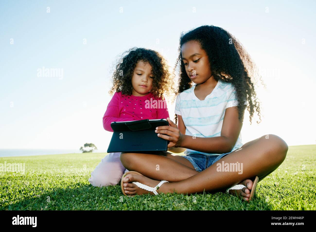 Junge gemischte Rasse Mädchen und ihre jüngere Schwester im Freien sitzen Teilen eines digitalen Tablets Stockfoto