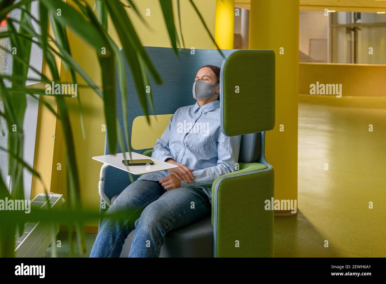 Frau in Gesichtsmaske schlafen entspannend auf bequemen Stuhl in der Ecke des Büros oder der Universität mit Telefon. Soziale Distanzierung, Isolation, Einsamkeit, Alon Stockfoto