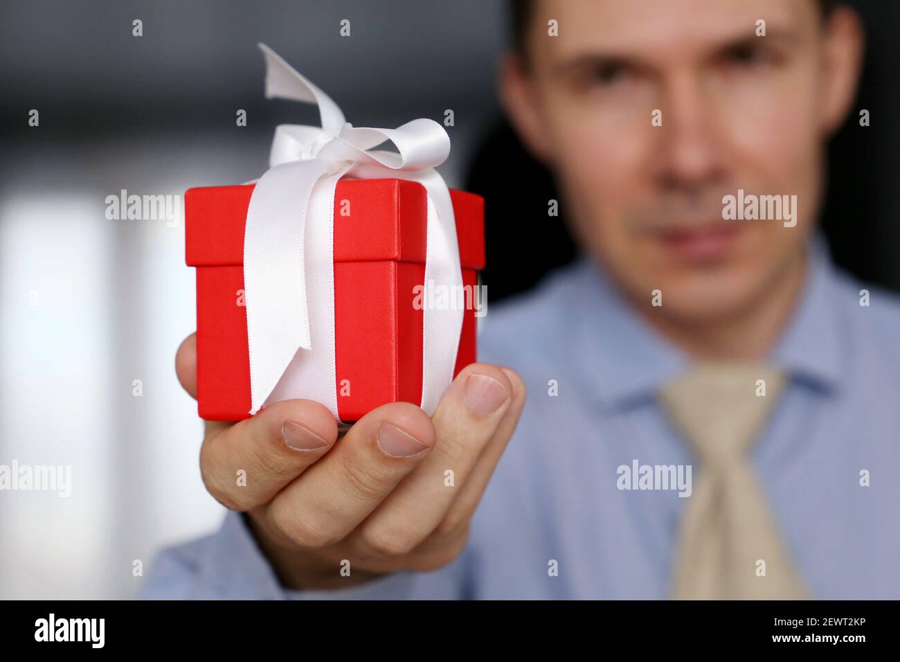 Rote Geschenkbox in männlicher Hand Nahaufnahme, Feiertagskonzept. Lächelnder Mann in Bürokleidung, selektiver Fokus auf Geschenk Stockfoto