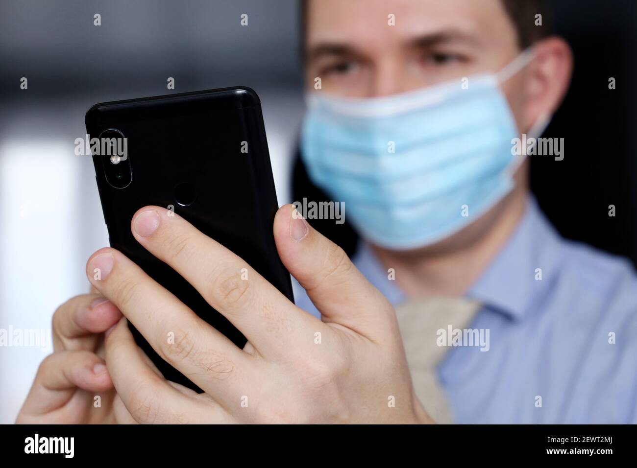 Smartphone in männlichen Händen Nahaufnahme, Mann in medizinischen Gesichtsmaske und Bürokleidung mit Handy. Kommunikation während einer Coronavirus-Pandemie Stockfoto