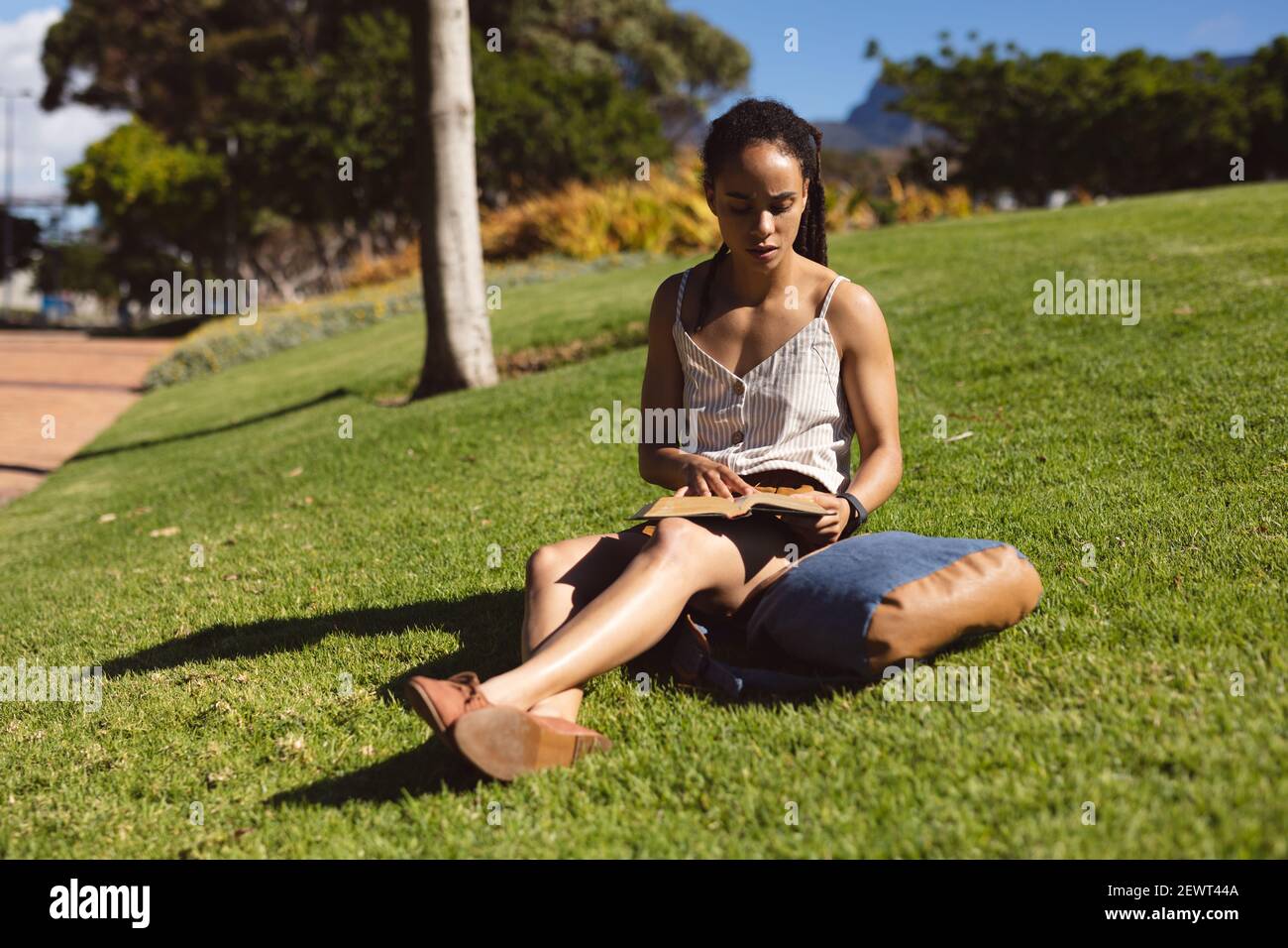 afroamerikanische Frau, die auf Gras sitzt, runzelte die Stirn und las im Park ein Buch Stockfoto