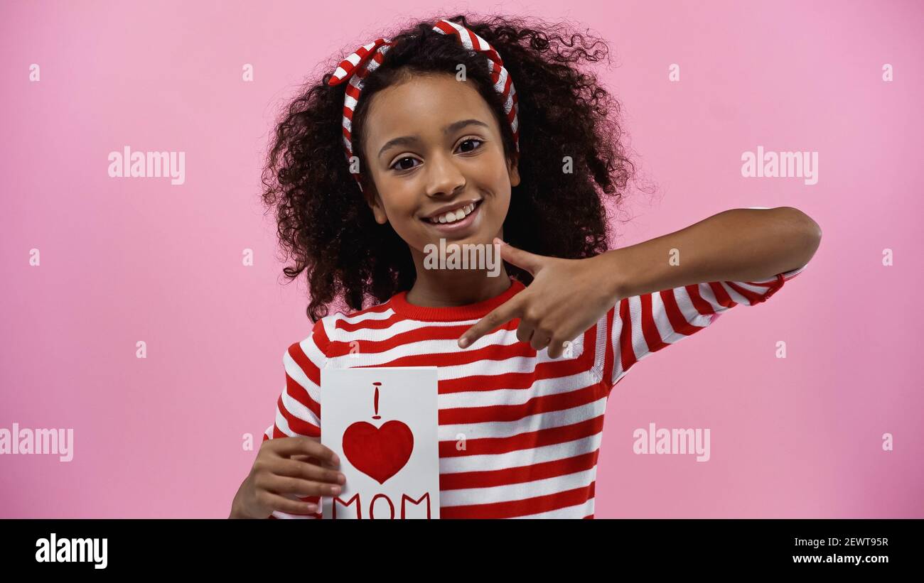 Happy african american Mädchen zeigt mit dem Finger auf Grußkarte Mit i love Mama Schriftzug isoliert auf rosa Stockfoto