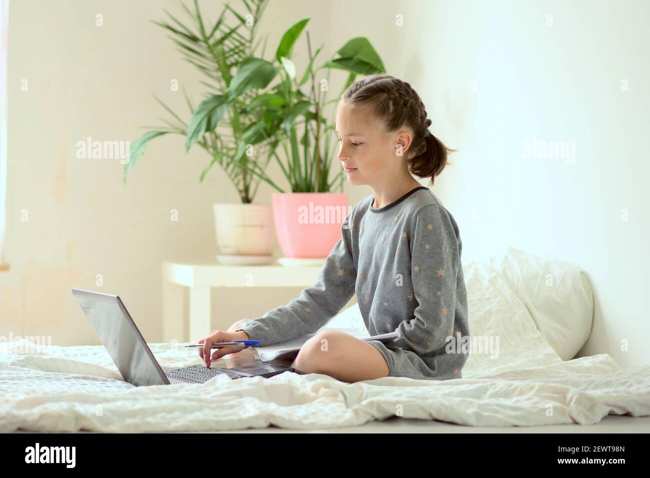 Eine junge Schülerin macht ihre Hausaufgaben und sitzt zu Hause auf dem Bett. Vor dem Mädchen ist ein Laptop. Sie hält einen Stift in der Hand. Selbstbildungs-Konzept. Nahaufnahme. Seitenansicht. Stockfoto