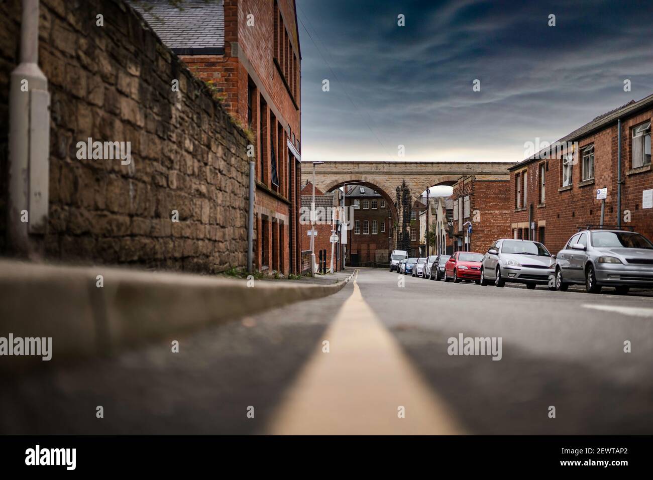 Keine Parkbeschränkungen Gelbe Linie auf der Straße niedrigen Winkel zeigt Autos geparkt geschäftigen Stadt Straße Szene. Eisenbahn Bögen Brücke Geschäfte und Häuser dramatischen Himmel Stockfoto
