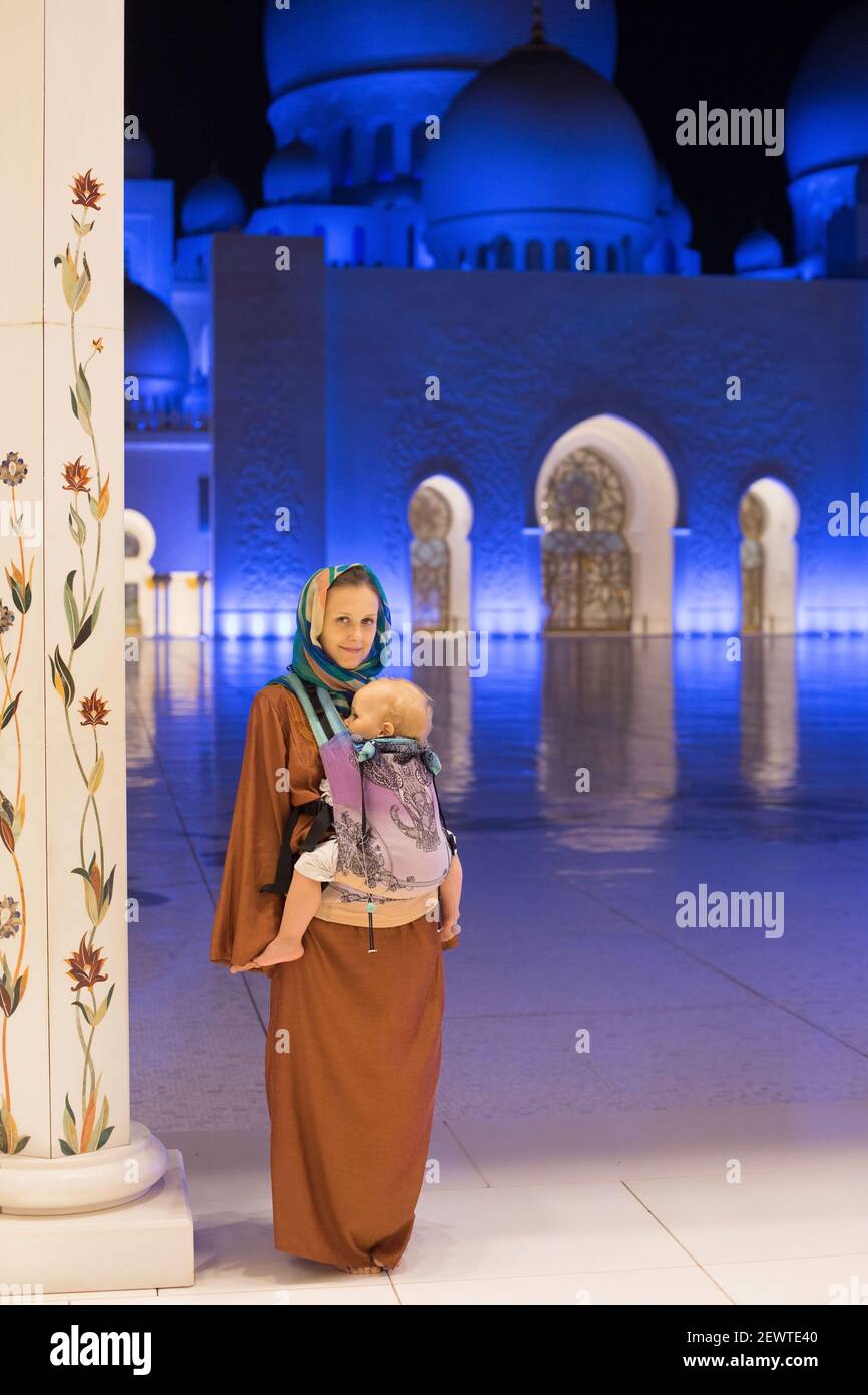 Junge Mutter, die mit Baby Junge in ergo Tasche Tragetuch, Aufenthalt in langen Kleid und Haarschutz in Grand weiße Moschee Hintergrund in Abu Dhabi, VAE Stockfoto