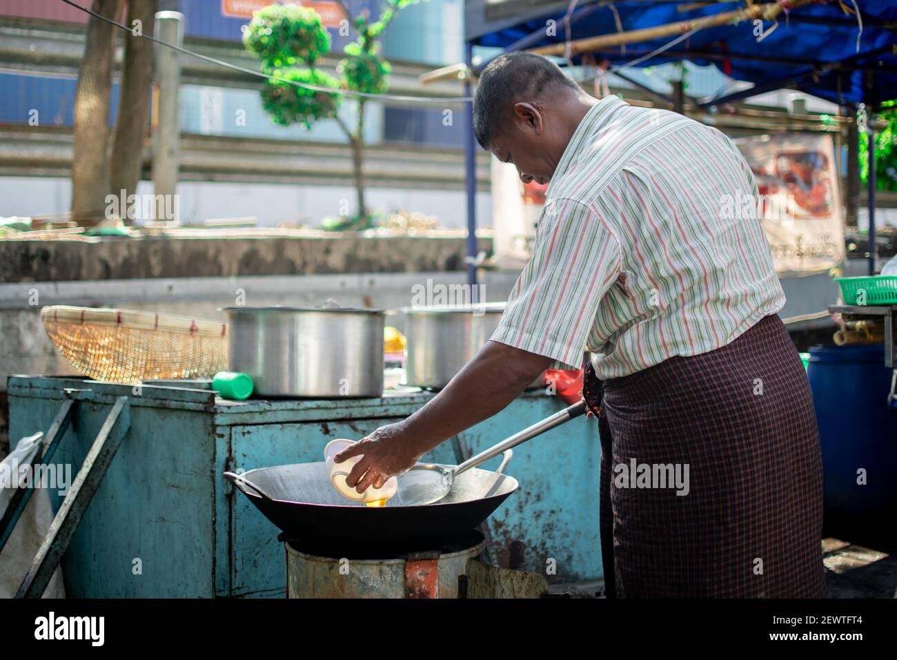 YANGON, MYANMAR - JANUAR 1 2020: Ein burmesischer Mann in traditionellem longyi frittiert ein Ei in einer Wok-Pfanne an einem kleinen Imbissstand in der Innenstadt Stockfoto