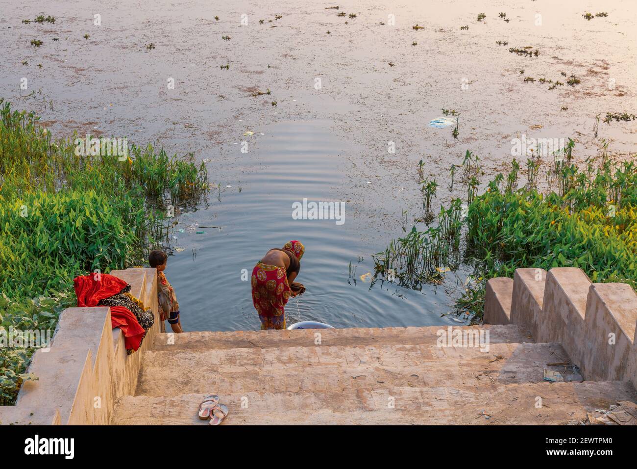 Ländliche Frau wäscht ihre Kleidung an einem Flussufer, während ihr Kind auf ein Dorf in Westbengalen, Indien schaut Stockfoto
