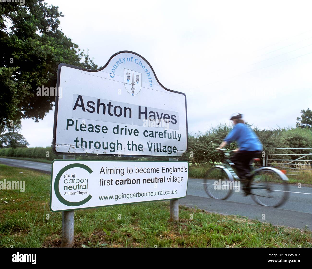 Fahrradfahrer, der am Schild "CO2-neutrales Dorf" vorbeifährt, Ashton Hayes, Cheshire. Stockfoto