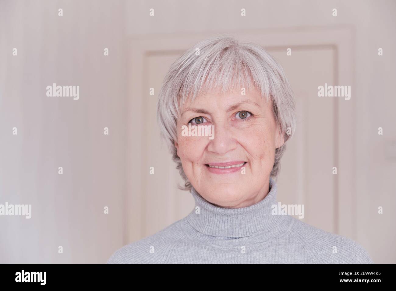 Ein Nahaufnahme Porträt einer lächelnden und Blick auf die Kamera ältere kaukasische Frau mit kurzen grauen Haaren auf einem hellen Hintergrund. Stockfoto