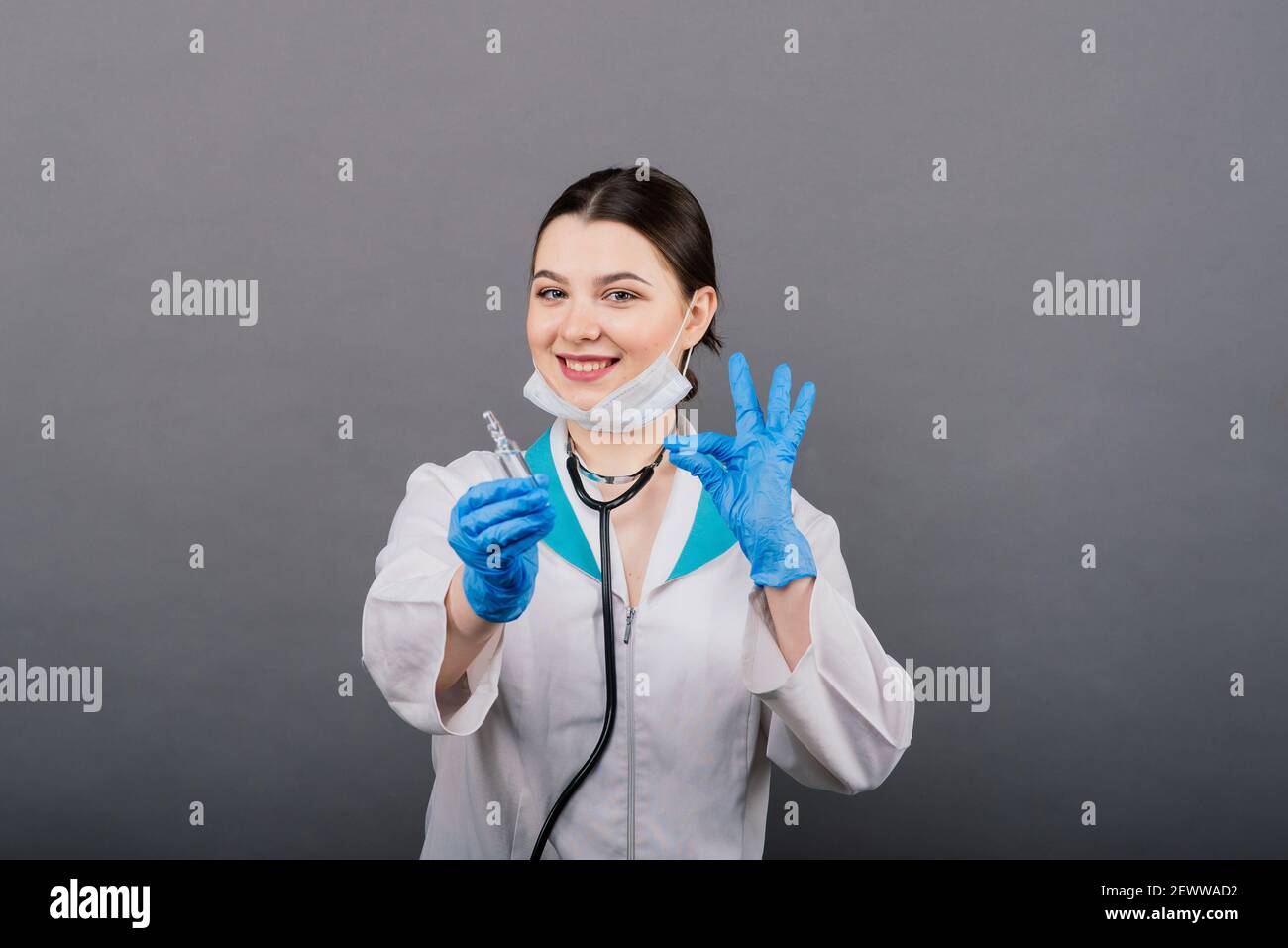 Seriöse Ärztin in einer medizinischen Maske hält Spritze und Impfstoff isoliert auf grau Stockfoto