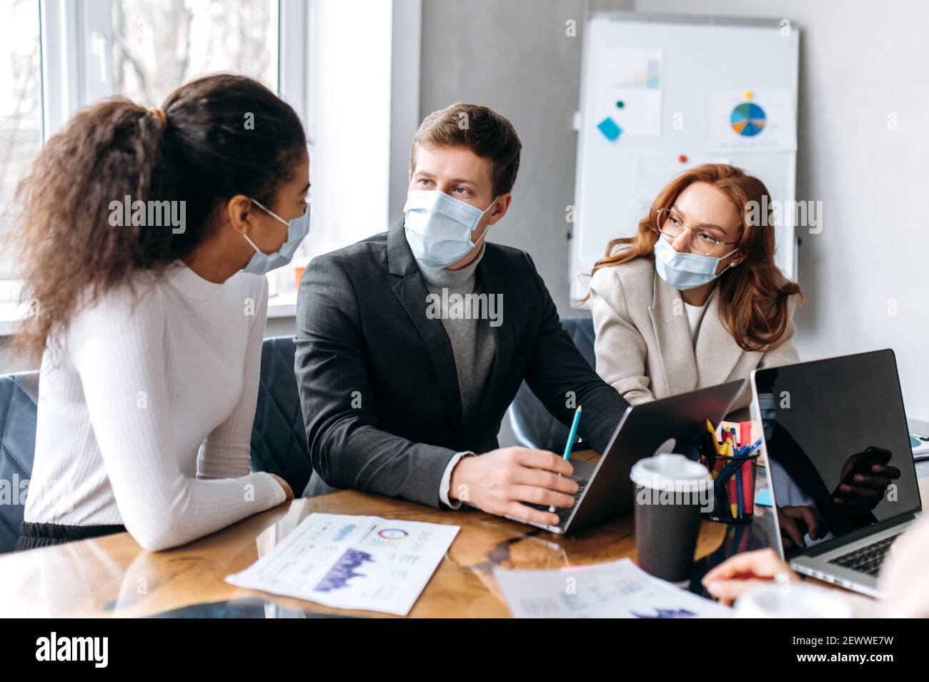 Konzentrierte Gruppe von Geschäftsleuten beim Briefing Meeting. Multirassische Mitarbeiter tragen medizinische Masken, studieren an Finanzgraphen, Brainstorming zusammen, diskutieren Ideen, arbeiten an neuen Projekt Stockfoto