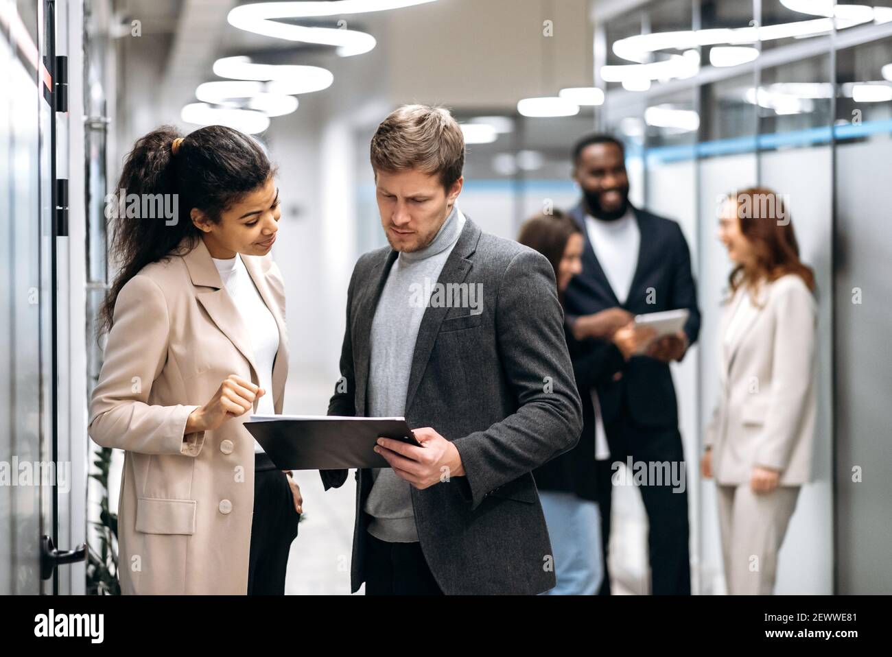 Seriöse multirassische Kollegen stehen in der Bürohalle, reden über Arbeitsfragen. Konzentrierte Geschäftspartner diskutieren Unternehmensprojekt, geben sich gegenseitig Ratschläge, Teamwork-Konzept Stockfoto
