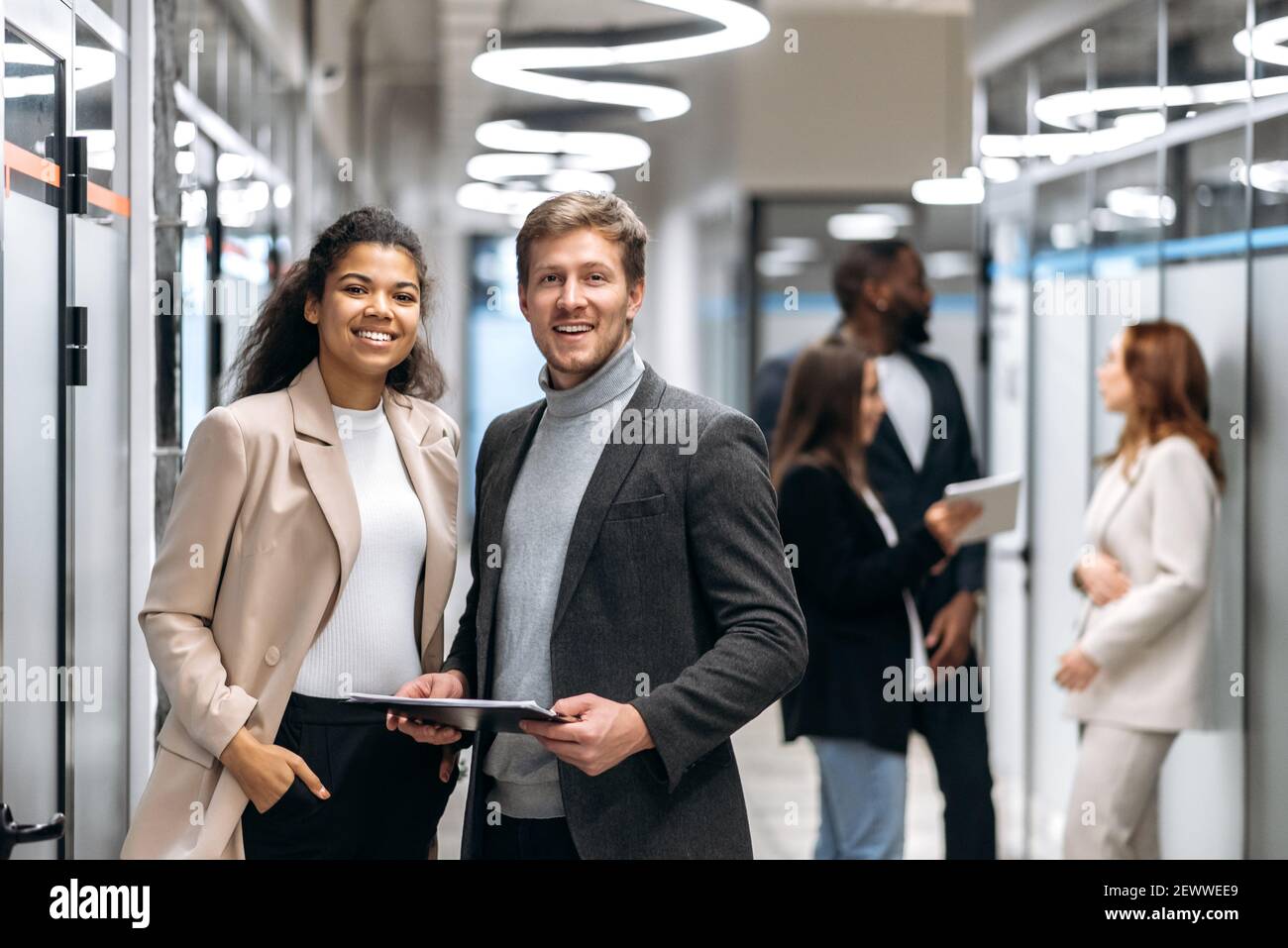 Porträt von zwei multirassischen zufriedenen erfolgreichen Kollegen in formeller Kleidung, die in einem modernen Büro stehen und freundlich lächeln auf die Kamera Stockfoto