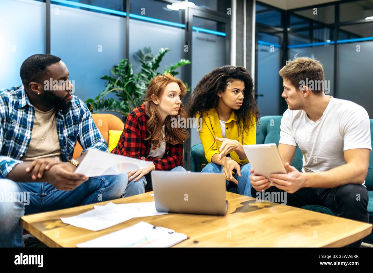 Konzentrierte Kollegen oder Studenten sitzen auf der Couch, diskutieren Arbeit oder Studienprojekt. Seriöse, multiethnische Kollegen arbeiten zusammen an der Präsentation oder am Finanzbericht Stockfoto