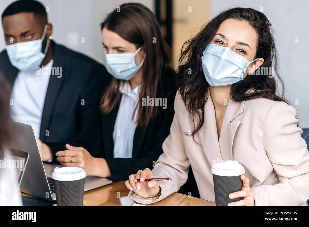 Gruppe von Geschäftsleuten bei Briefing Meeting. Multirassische Kollegen tragen medizinische Masken, Brainstorming zusammen, diskutieren Ideen, arbeiten an neuen Projekt, eine Frau schaut auf Kamera Stockfoto
