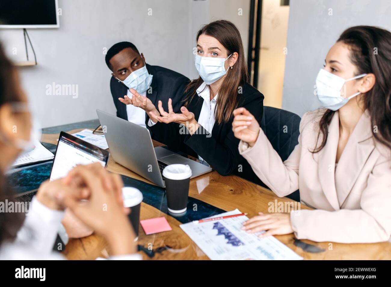 Konzentrierte Gruppe von Geschäftsleuten beim Briefing Meeting. Multirassische Mitarbeiter tragen medizinische Masken, studieren an Finanzgraphen, Brainstorming zusammen, diskutieren Ideen, arbeiten an neuen Projekt Stockfoto
