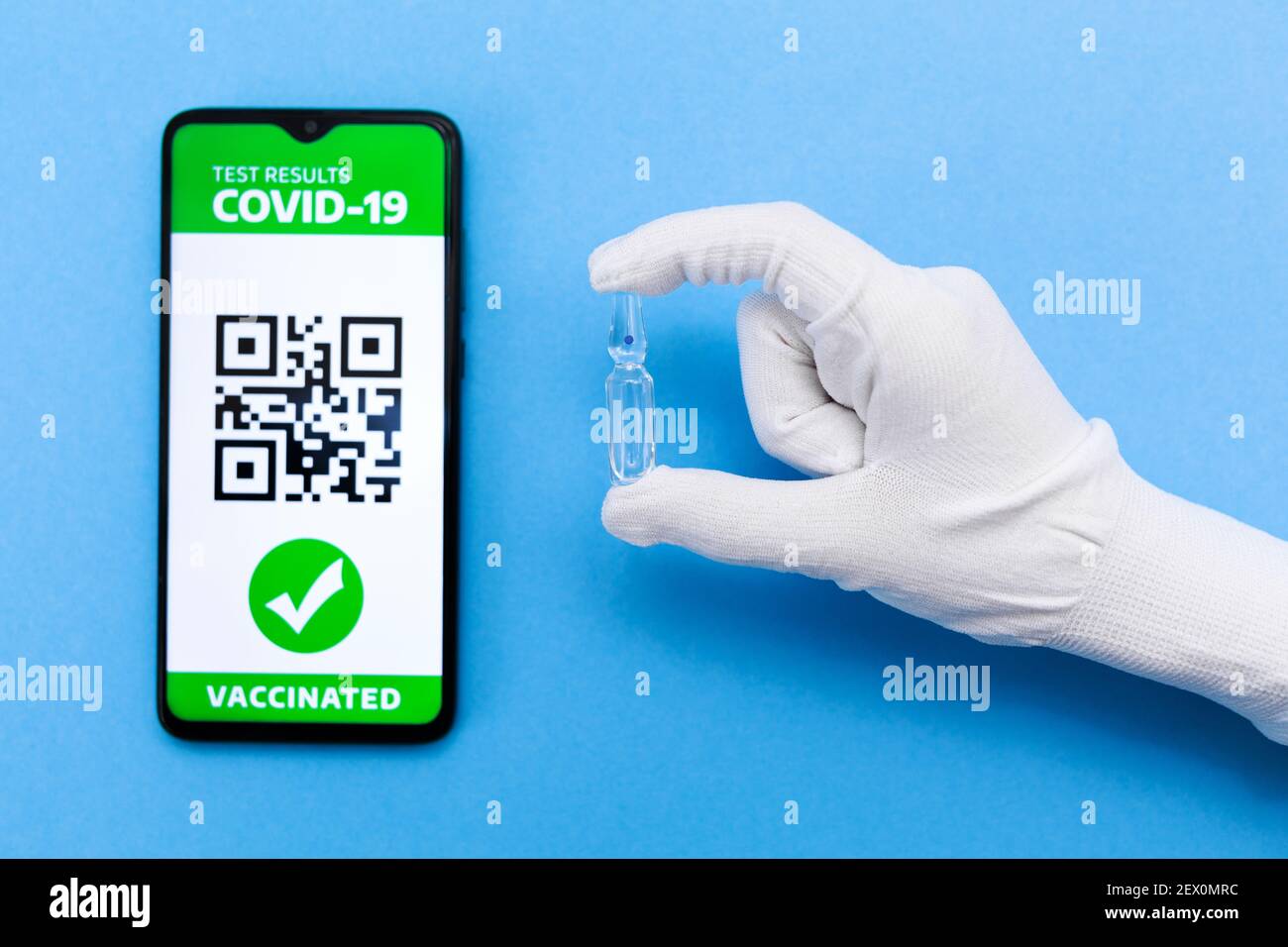 Eine Hand im Handschuh hält eine Impfampulle. Handy-Bildschirm mit einem elektronischen Pass der Immunität mit COVID-19 Impfung. Blauer Hintergrund. Flach Stockfoto