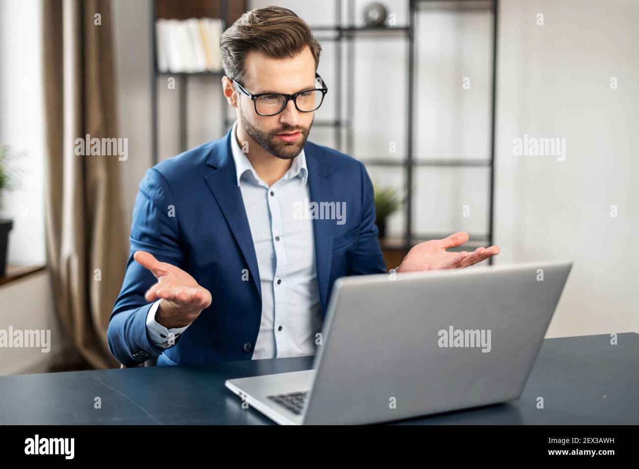 Clever gepflegter Geschäftsmann mit stylischen Brillen sitzt auf seinem Arbeitsplatz im modernen Büro, sieht auf Laptop-Bildschirm, zeigt mit Händen und spricht online. Remote-Arbeit, Arbeit in der Ferne Stockfoto