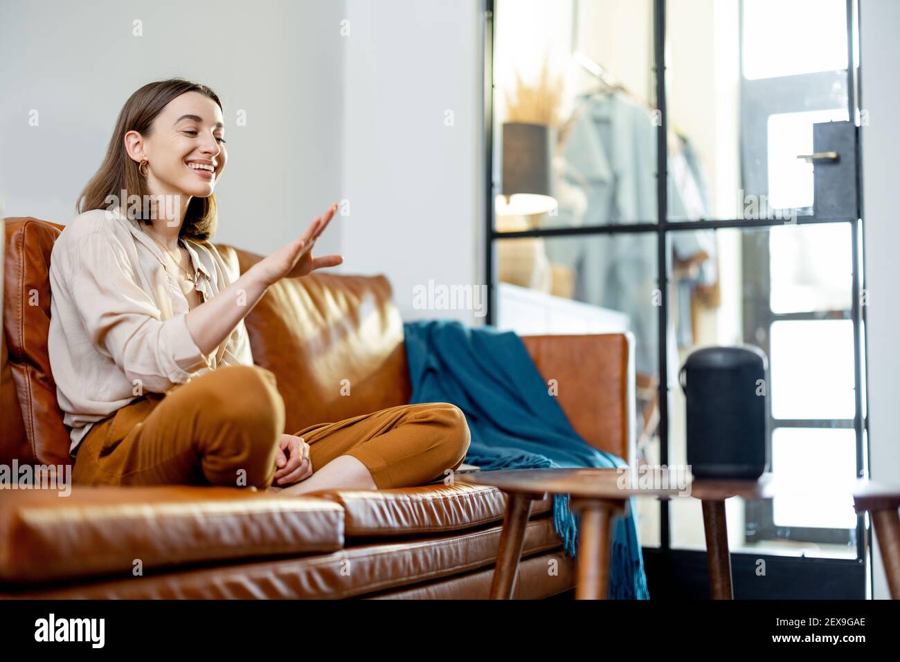 Glückliche hübsche Frau, die mit schwarzem Audio-Assistent Spalte auf dem Couchtisch spricht, während sie auf braunem Ledersofa im stilvollen Wohnzimmer sitzt. Smart Home-Konzept. Stockfoto