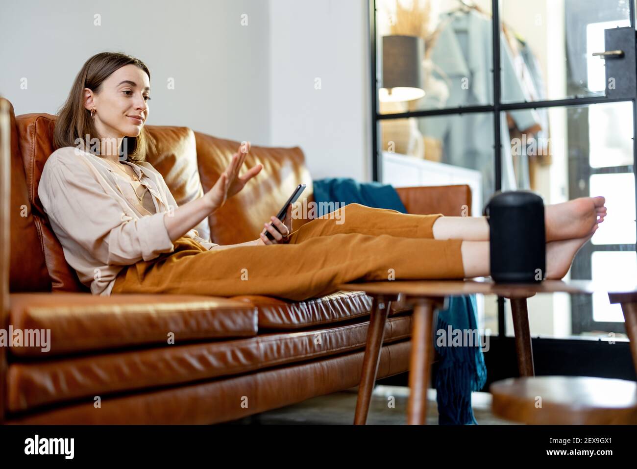 Glückliche hübsche Frau, die mit schwarzem Audio-Assistent Spalte auf dem Couchtisch spricht, während sie auf braunem Ledersofa im stilvollen Wohnzimmer sitzt. Smart Home-Konzept. Stockfoto