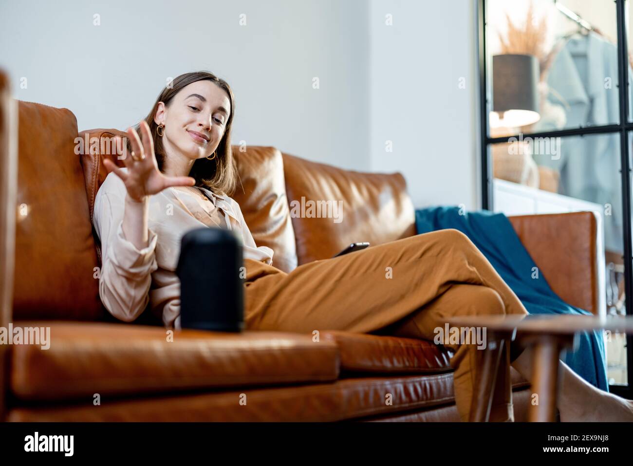Glückliche hübsche Frau, die mit schwarzem Audio-Assistent Spalte auf dem Couchtisch spricht, während sie auf braunem Ledersofa im stilvollen Wohnzimmer sitzt. Smart Home-Konzept. Stockfoto