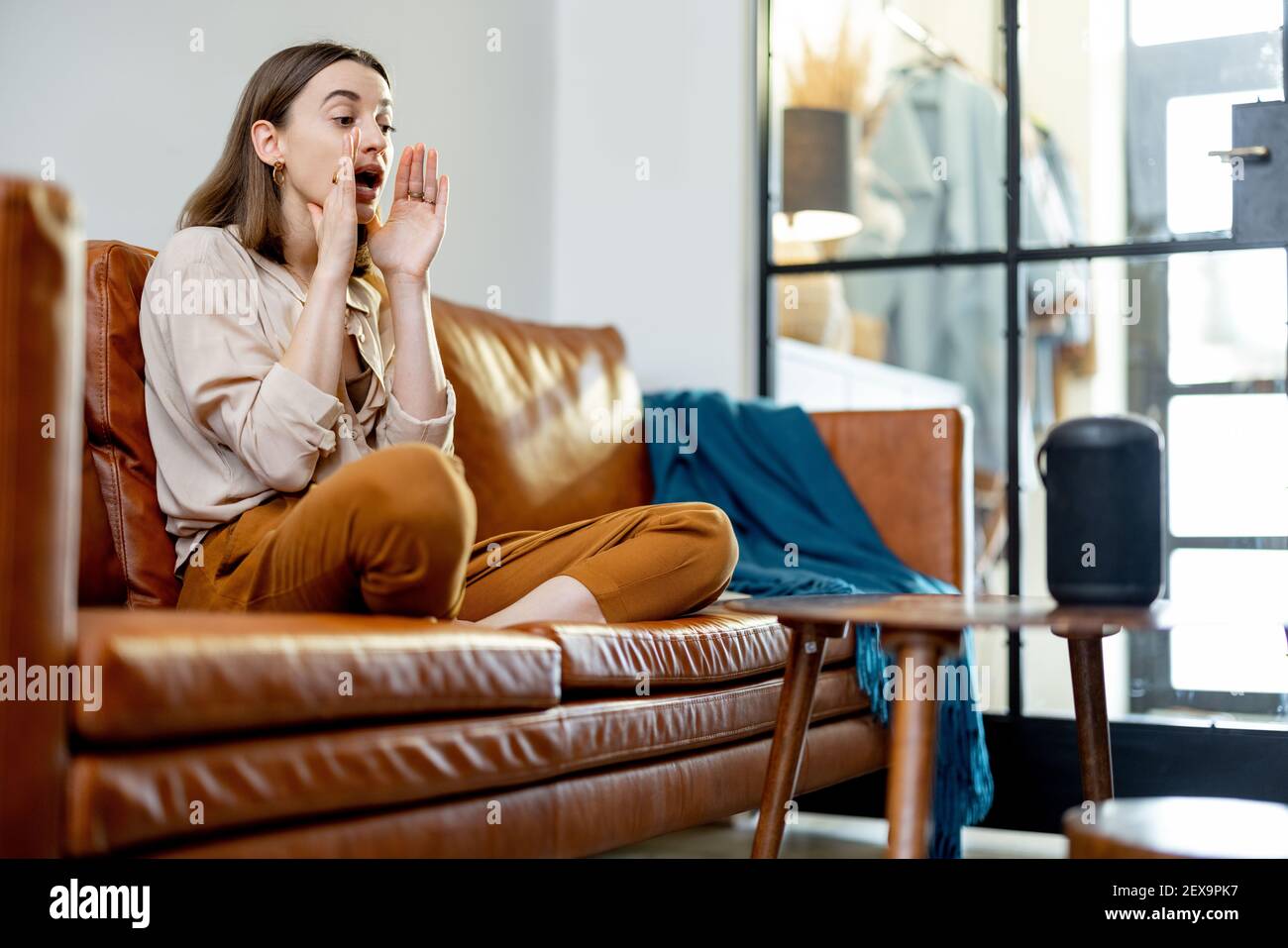 Glückliche hübsche Frau, die mit schwarzem Audio-Assistent Spalte auf dem Couchtisch spricht, während sie auf braunem Ledersofa im stilvollen Wohnzimmer sitzt. Smart Home-Konzept. Stockfoto