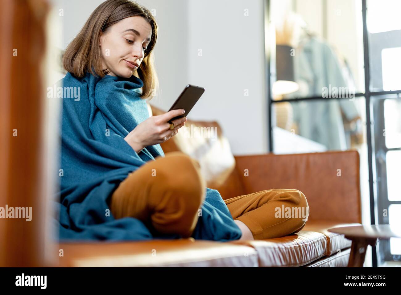 Hübsche Frau mit Smartphone bedeckt mit einem blauen karierten Sitz auf dem braunen Sofa zu Hause. Lifestyle und Wohnlichkeit Konzept. Stockfoto