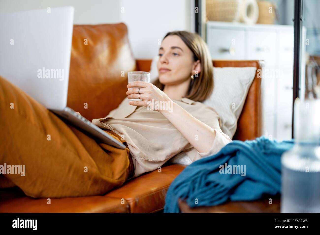 Hübsche Frau hält ein Glas Wasser, während sie am Laptop arbeitet und auf dem Sofa liegt. Lifestyle und gesundes Konzept. Austrocknung. Trinkwasserquote pro Tag Stockfoto