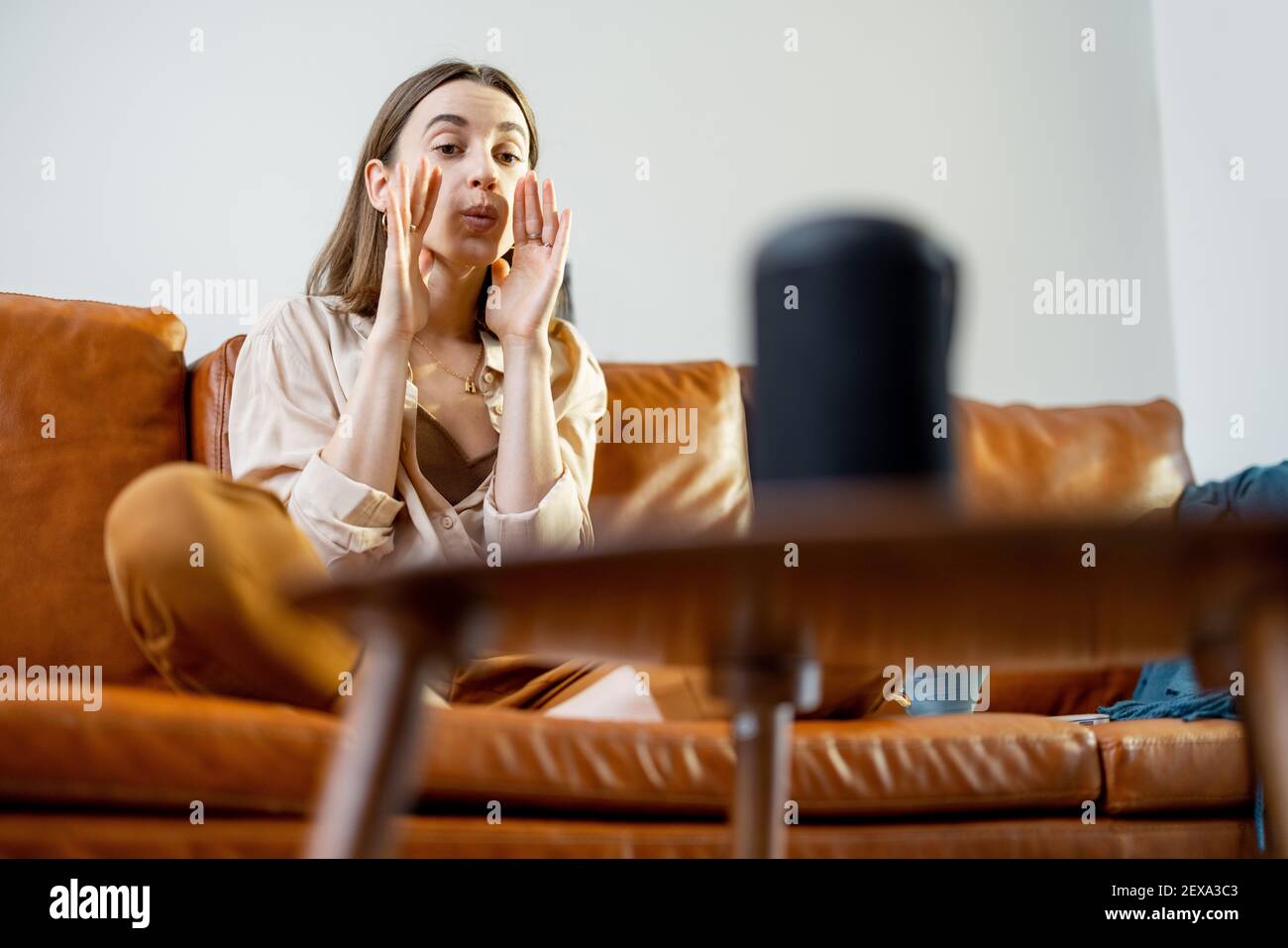 Glückliche hübsche Frau, die mit schwarzem Audio-Assistent Spalte auf dem Couchtisch spricht, während sie auf braunem Ledersofa im stilvollen Wohnzimmer sitzt. Smart Home-Konzept. Stockfoto