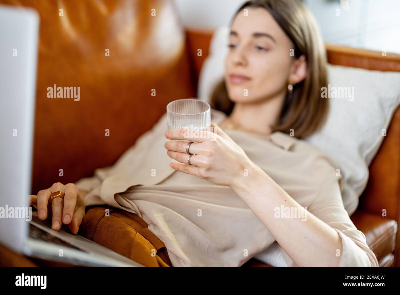 Hübsche Frau hält ein Glas Wasser, während sie am Laptop arbeitet und auf dem Sofa liegt. Lifestyle und gesundes Konzept. Austrocknung. Trinkwasserquote pro Tag. Konzentrieren Sie sich auf das Glas. Stockfoto
