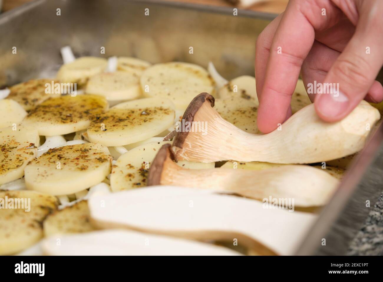 Nahaufnahme von Menschenhänden, die in Scheiben geschnittene Trompetenpilze (pleurotus eryngii) über gewürzte Kartoffeln und Zwiebeln legen. Kochvorgang. Mediterrane Ernährung. Stockfoto