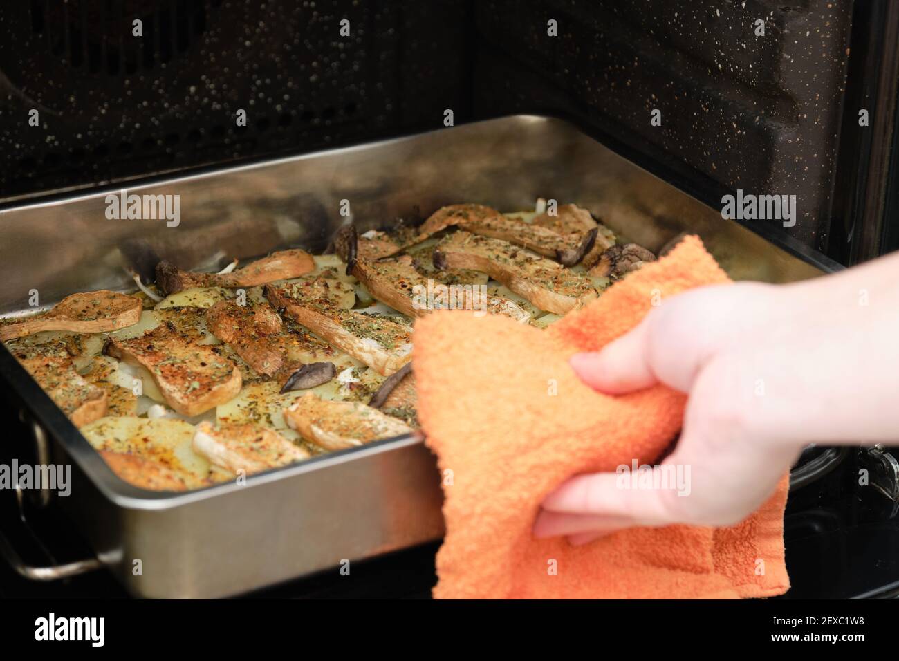 Zugeschnittenes Bild von Menschenhänden, die ein Tablett mit gewürzten, in Scheiben geschnittenen Trompetenpilzen (pleurotus eryngii), Kartoffeln und Zwiebeln aus dem Ofen nehmen. Mittelmeer Stockfoto