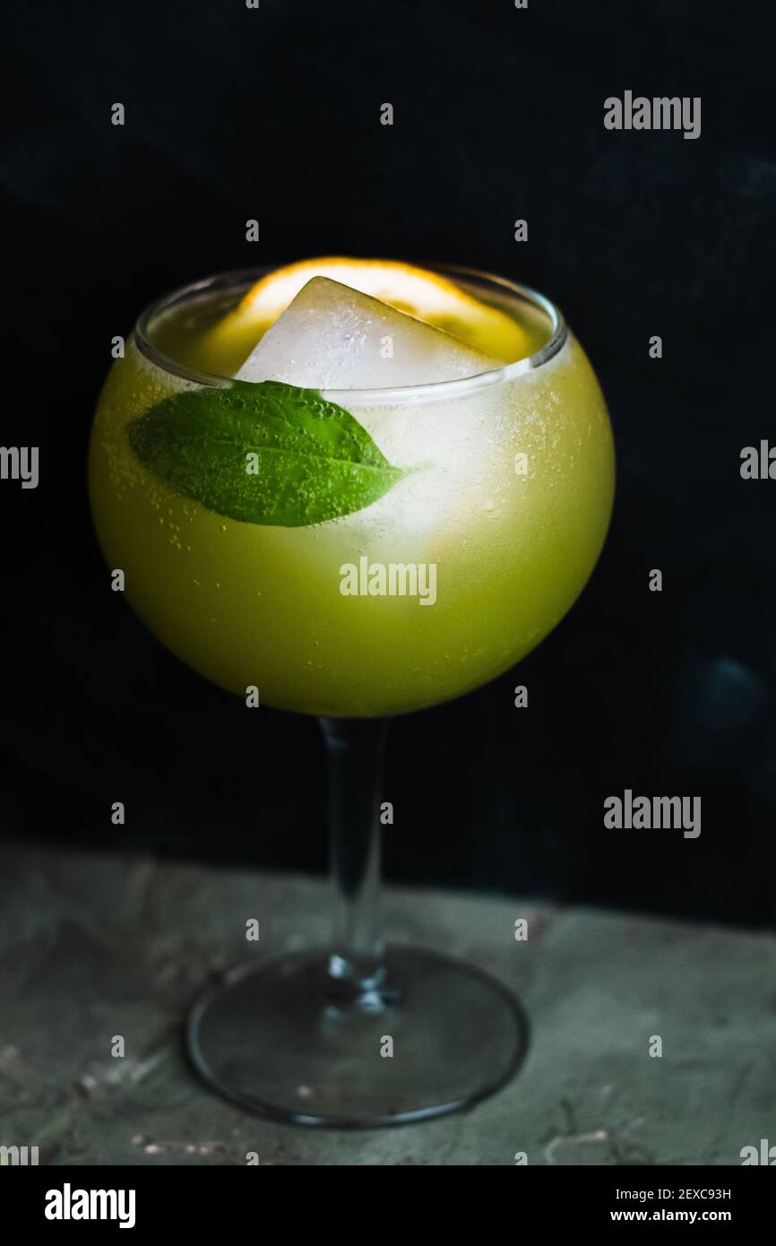 Helles grünes Getränk in einem Kelchglas mit einem großen quadratischen Eiswürfel, Zitronenscheibe und einem einzigen Basilikumblatt. Stockfoto