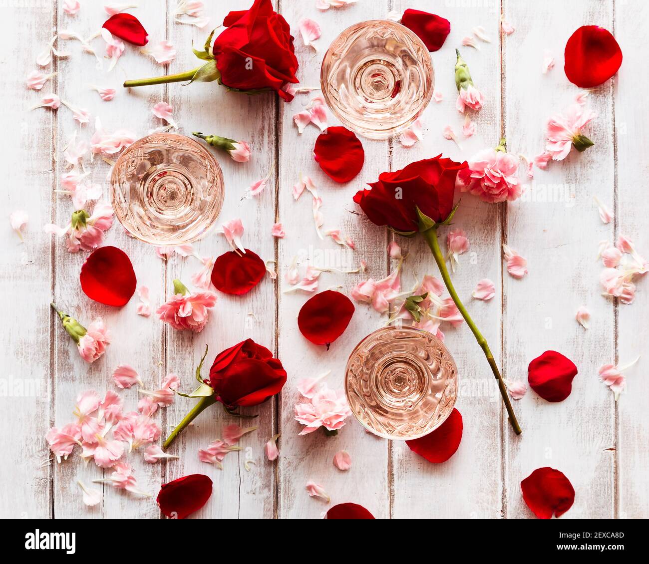 Rosé gefüllte Vintage-Coupé-Gläser auf einer weiß getünchten Holzoberfläche, umgeben von roten und rosa Blüten und Blütenblättern. Stockfoto