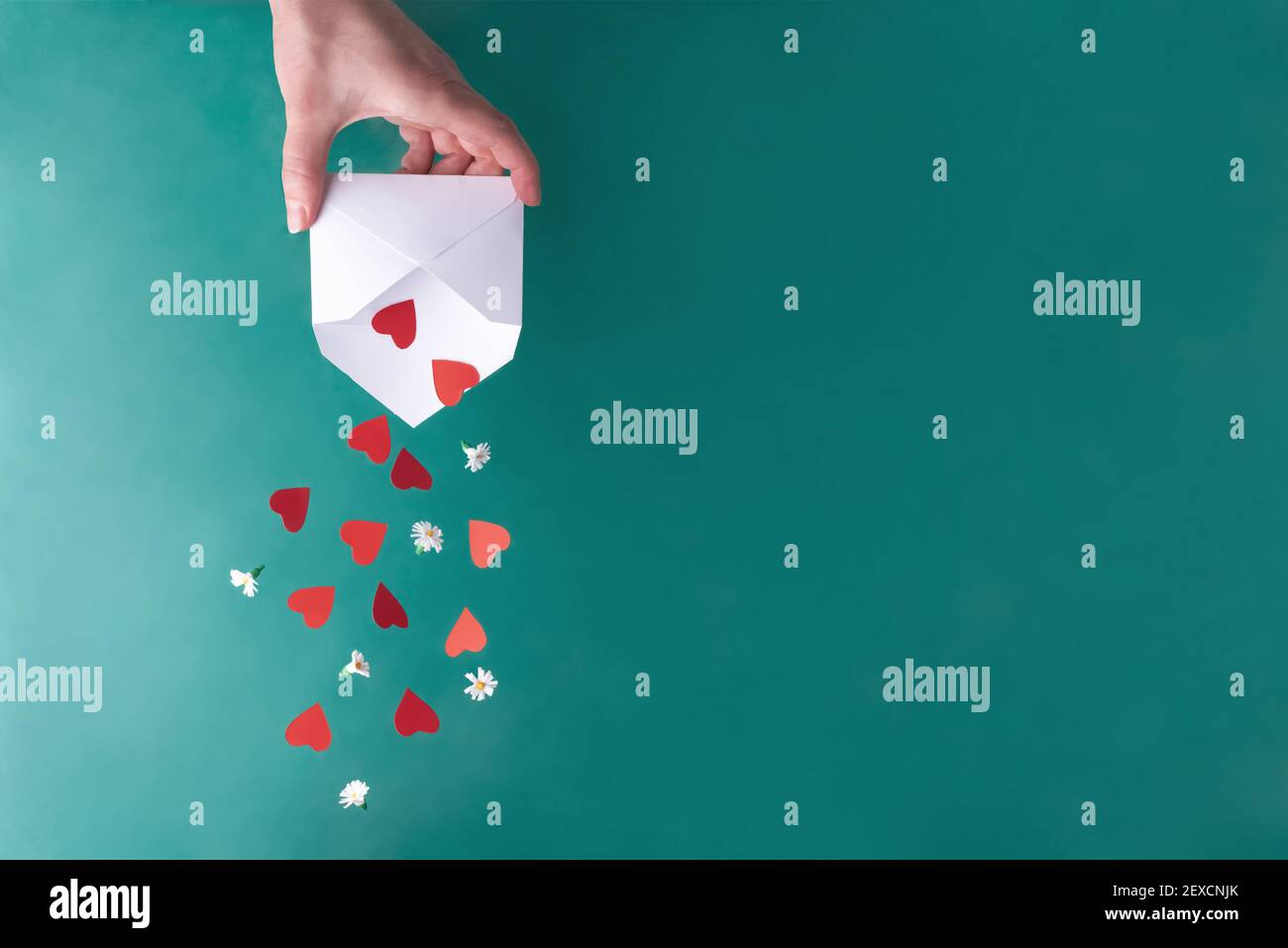 Weibliche Hand schüttelt rote Herzen und weiße Blumen aus einem weißen Umschlag auf einem türkisfarbenen Hintergrund, Nahaufnahme, Kopierraum. Valentinskarte. Stockfoto