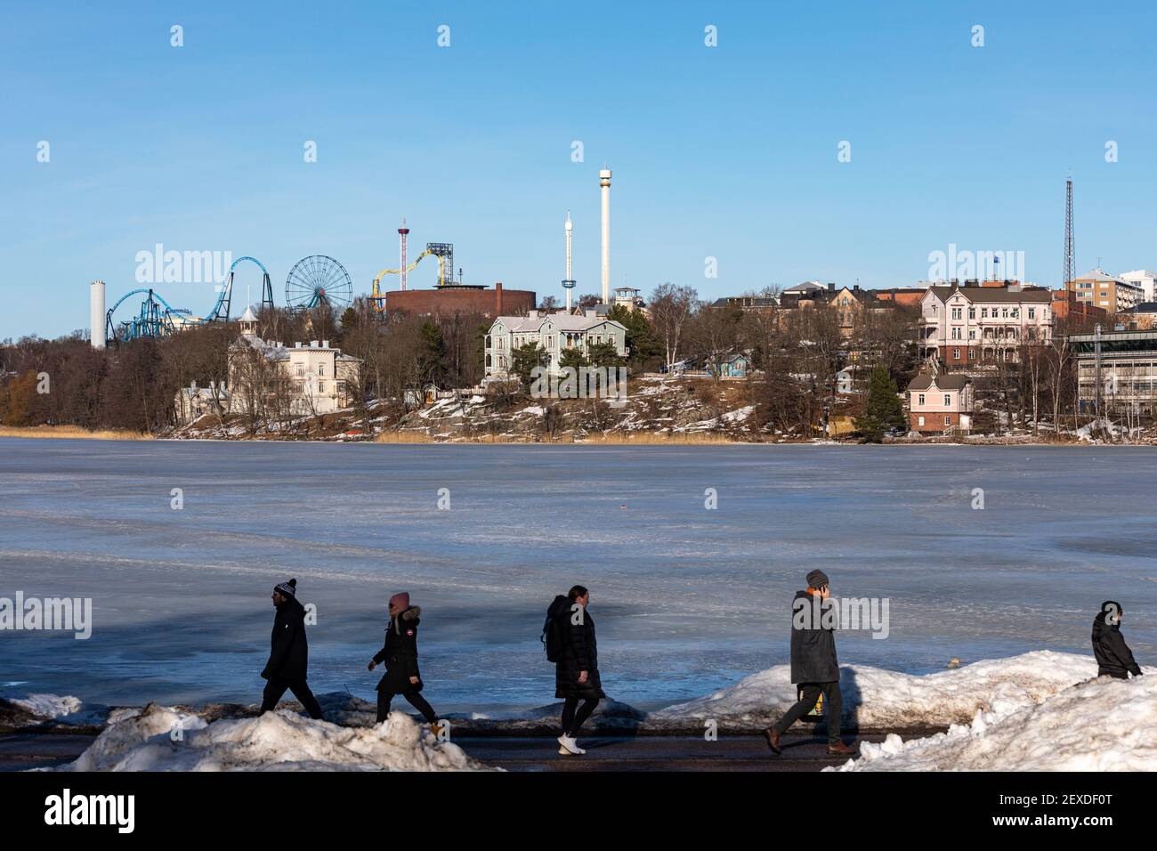 Gefrorene Töölönlahti Bucht mit vorbeifahrenden Menschen vor und Linnunlaulu Villen und Linnanmäki Vergnügungsfahrten im Hintergrund in Helsinki, Finnland Stockfoto