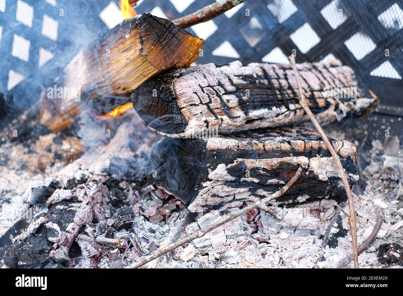 Ein Feuer in einem Metallkamin brennt draußen, mit weißer, glühender heißer Glut der Kohle aus brennendem Holz. Rauch steigt aus den verkohlten Baumstämmen und Stockfoto