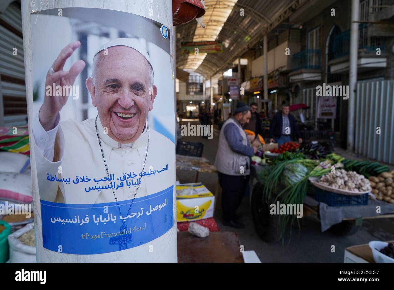 Ein Plakat von Papst Franziskus auf einem Pfeiler neben einem Gemüsehändler.Jugendliche Iraker haben eine Kampagne gestartet, um Plakate von Papst Franziskus in den Straßen der Altstadt der Stadt Mosul zu verbreiten, Begrüßung seines Besuchs in der Stadt am 7. März 2021 während des historischen Besuchs im Irak vom 5. Bis 8. März. Sein historischer Besuch beinhaltet eine Reise in mehrere Städte wie die Hauptstadt Bagdad, die Stadt Mosul im Norden und ein Treffen mit der schiitischen religiösen Autorität Ayatollah Ali Sistani. Stockfoto