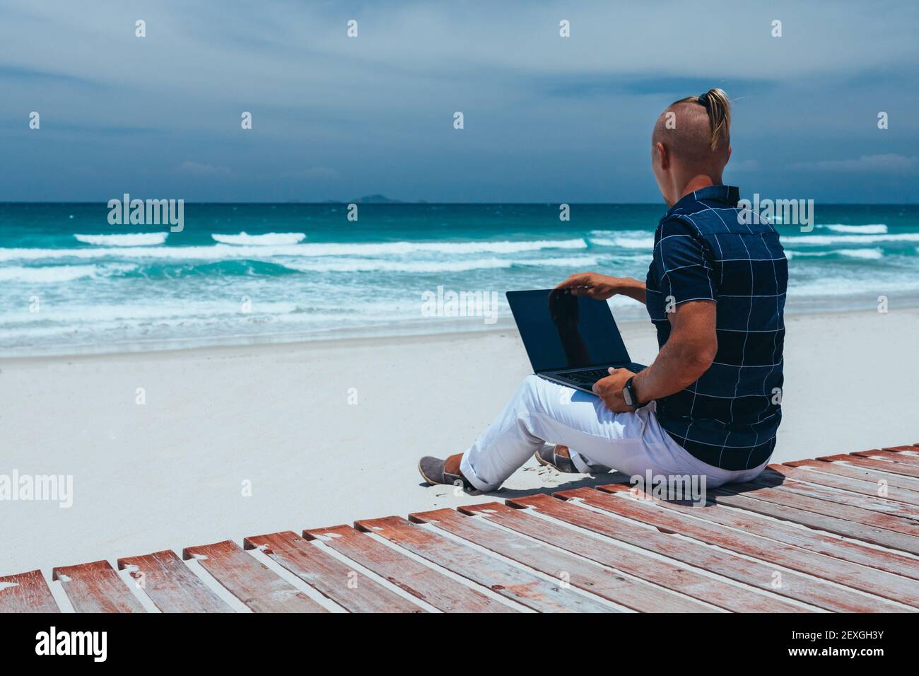 Guy öffnet einen Laptop für die Arbeit, sitzt am Strand an einem Sandstrand. Geschäftsmann, der auf einer Reise an Remote-Arbeit arbeitet. Blogger, Freiberufler macht einen Co Stockfoto