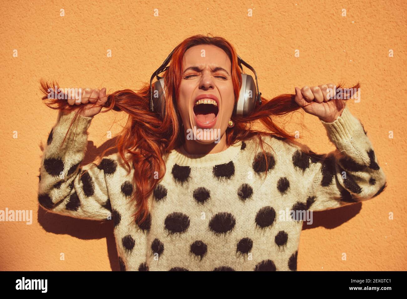 Weiße kaukasische Frau mit roten Haaren trägt Helme, um sich vor Lärm zu schützen zieht ihre Zöpfe beim Schreien. Geräuschkonzept Stockfoto