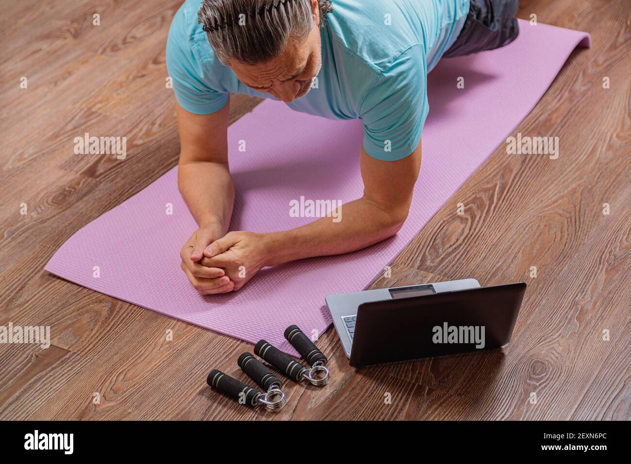 Der 50-jährige Mann führt Übungen durch, während er zu Hause auf dem Teppich liegt und den Computer anschaut. Während einer Pandemie trainiert eine Person in einer Wohnung über den Stockfoto