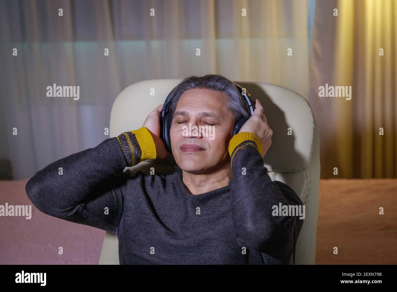 Der 50-jährige Mann hört zu Hause Musik auf Kopfhörern, sitzt auf einem Stuhl. Erholsame Freude. Stockfoto