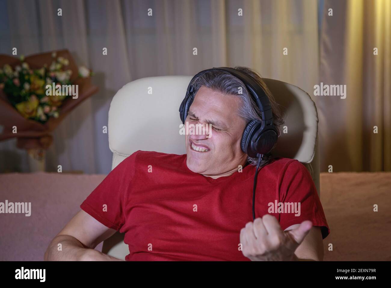 Der 50-jährige Mann hört zu Hause Musik auf Kopfhörern, sitzt auf einem Stuhl. Erholsame Freude. Stockfoto