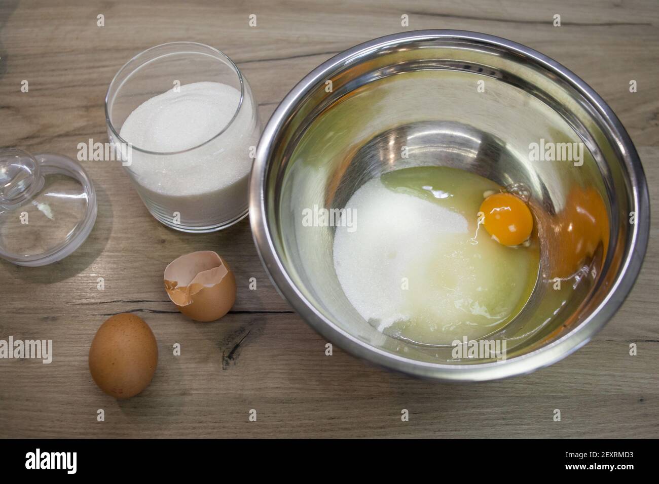Weißer Zucker und Ei in einer Schüssel und zwei Eier auf dem Holzhintergrund. Der zweite Schritt für die Herstellung von Muffins, Kuchen, Keks. Backvorgang Schritt für Schritt. Stockfoto