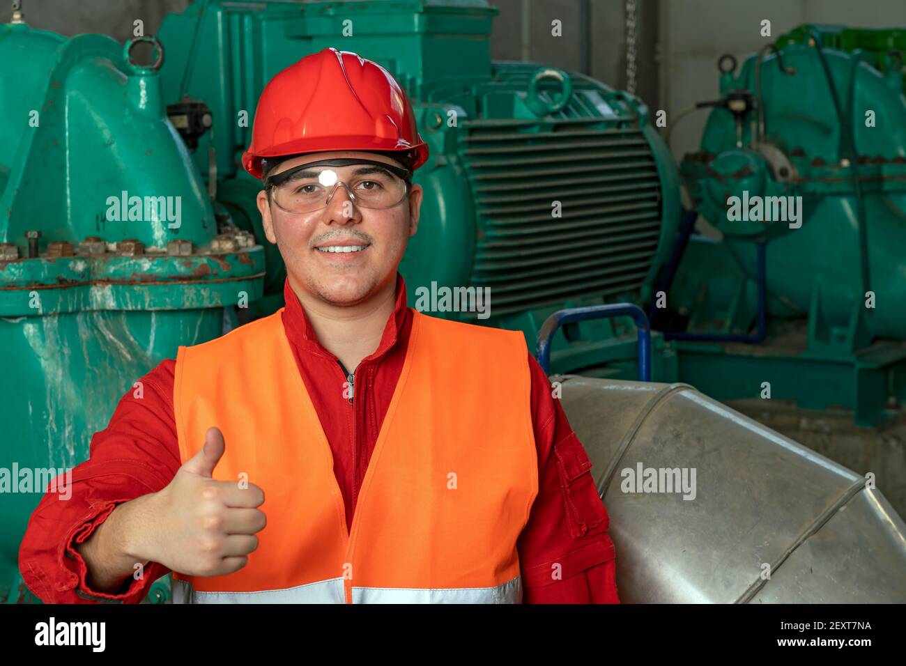 Porträt eines lächelnden jungen Arbeiters mit Daumen nach oben im Fernwärmekraftwerk. Arbeiter in persönlicher Schutzausrüstung, der die Kamera betrachtet. Stockfoto