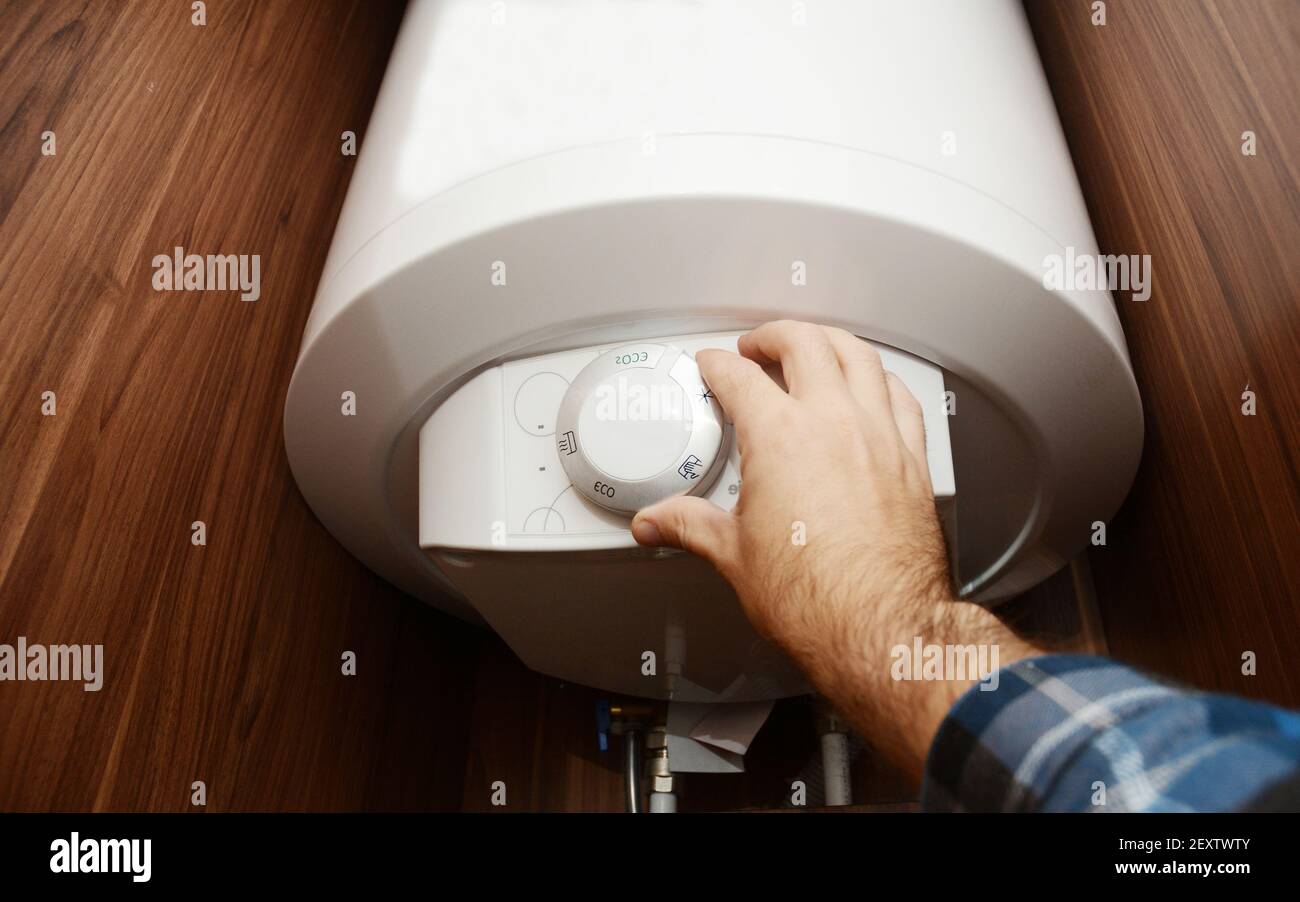 Ein Mann schaltet auf einem elektrischen Warmwasserboiler, Warmwasserbereiter in einem Badezimmerschrank Einstellung der Temperatur des Heißwassers für Kessel eff Stockfoto
