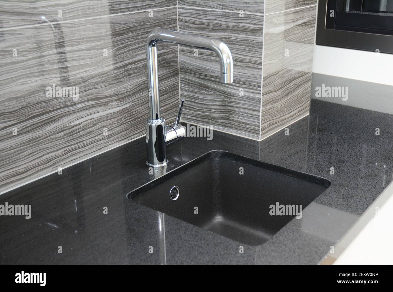 Eine Nahaufnahme einer modernen Küche, die mit grauen Keramikfliesen mit geschwungenem Liniendesign, schwarzer Küchenarbeitsplatz und quadratischem, schwarzem Unterbodenwaschbecken gefliest ist. Stockfoto