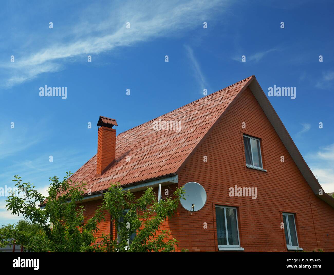 Ein Haus aus einfacher Bauweise aus roten Ziegeln mit einem Metalldach, einem Kamin, Dachrinnen, einem Dachfenster und einer Satellitenantenne. Stockfoto
